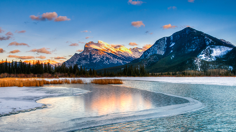 Snowcapped mountains backing a nearly frozen body of water with green trees along the shores