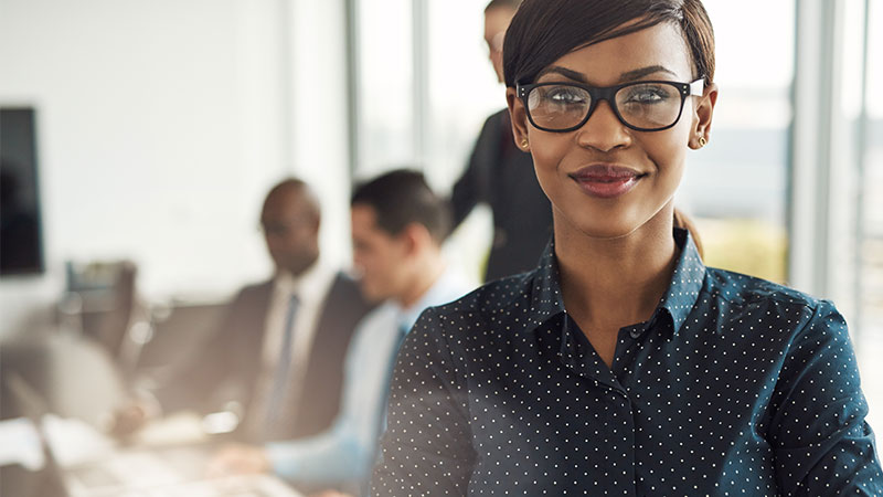 Person smiling with three other people in the background in an office  working at a computer