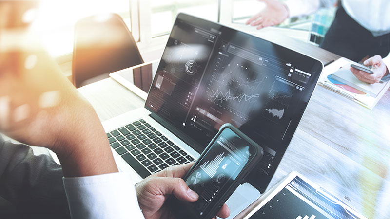 Close up of someone using a laptop and a phone displaying data 